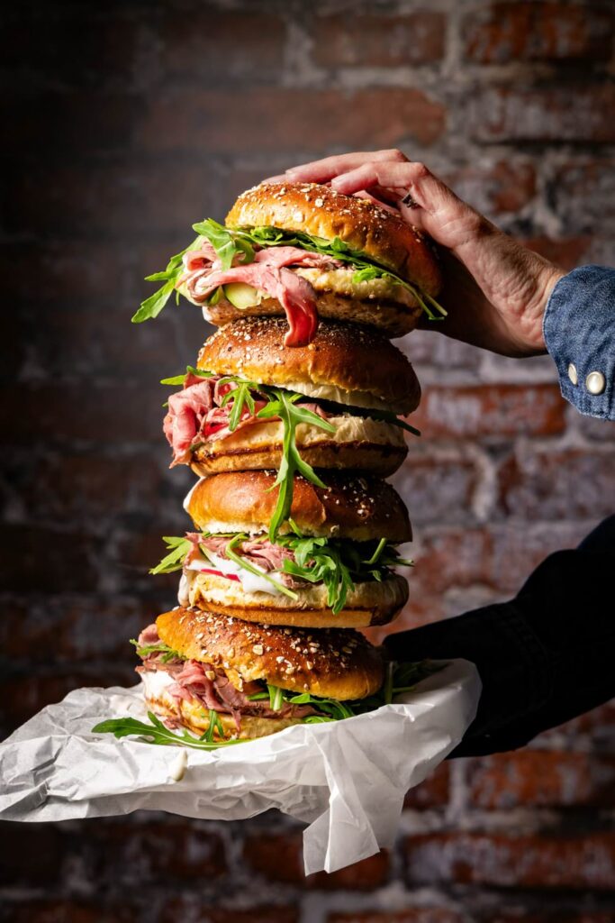 Gourmet Roastbeef Sandwich mit Rucola und Sesam-Bun – Food-Fotografie eines gestapelten Deli-Burgers im rustikalen Ambiente mit Brick-Wall Hintergrund