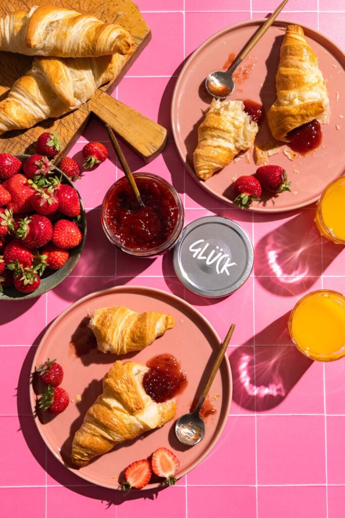 Modern inszeniertes Food-Fotografie-Bild mit Croissants, Erdbeermarmelade und frischen Erdbeeren auf pinkem Hintergrund – kreative Produktfotografie für Food Brands und Gastronomie