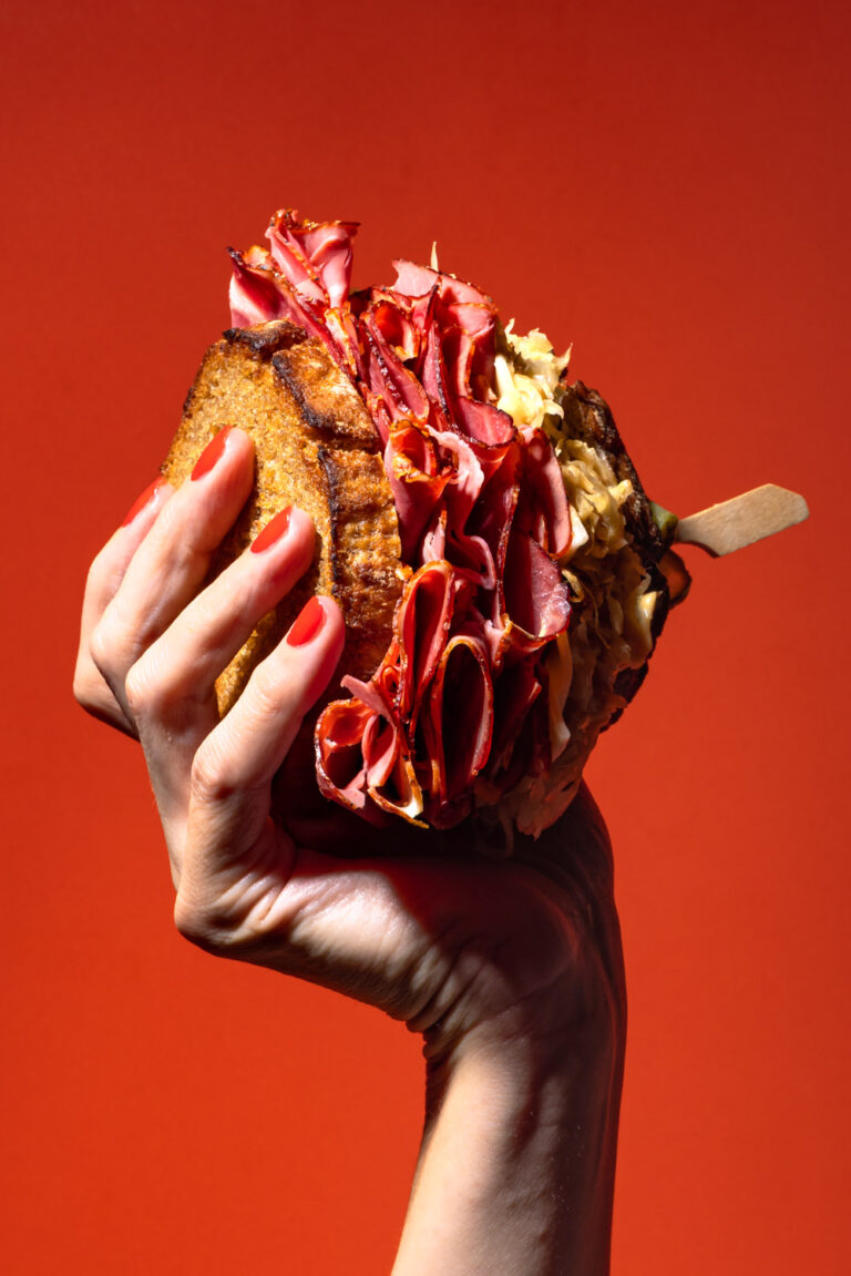 Reuben Sandwich: Pastrami-Sandwich mit Sauerkraut auf geröstetem Roggentoast, gehalten von einer Hand mit rot lackierten Fingernägeln vor rotem Hintergrund – professionelle, moderne Food-Fotografie